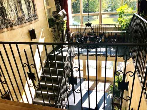 A bright staircase with a wrought iron railing, featuring a chandelier and a statue, surrounded by large windows and greenery.