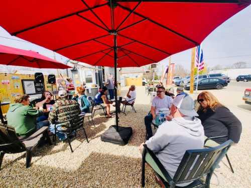 A lively outdoor gathering under red umbrellas, with people chatting and enjoying drinks in a gravel area.