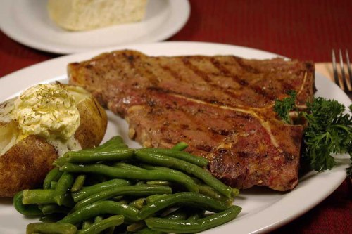 A plate featuring a grilled steak, baked potato with sour cream, and green beans, with a slice of bread in the background.