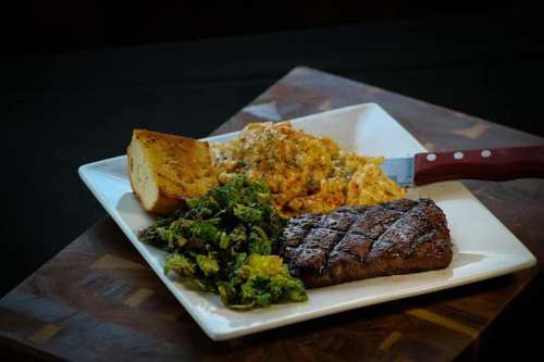 A plate featuring grilled steak, creamy pasta, sautéed greens, and a slice of cornbread on a wooden cutting board.