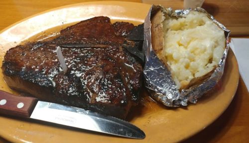 A juicy steak on a plate with a baked potato wrapped in foil, accompanied by a knife and fork.