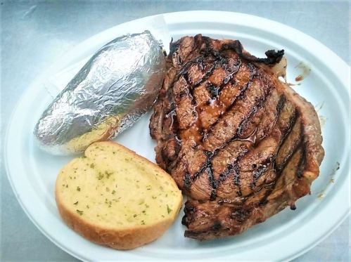 A grilled steak with a baked potato wrapped in foil and a slice of garlic bread on a white plate.
