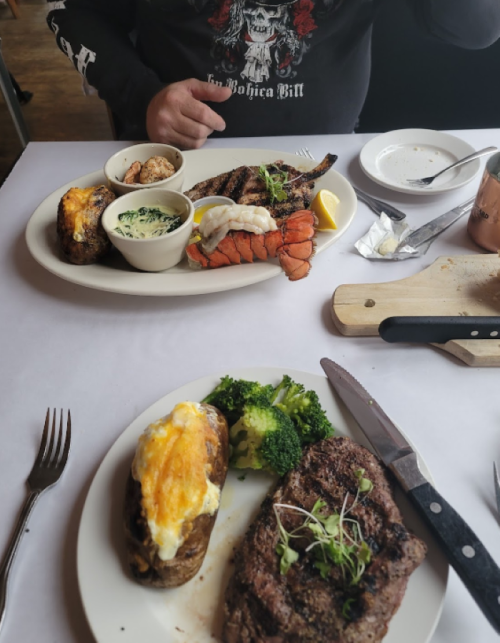A table setting with two plates: one with lobster, baked potato, and spinach; the other with steak and broccoli.