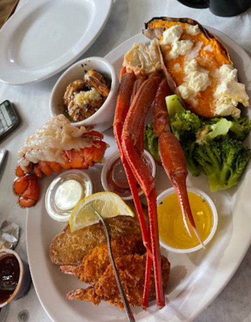A plate of seafood featuring lobster, crab legs, shrimp, baked sweet potato, broccoli, and dipping sauces.