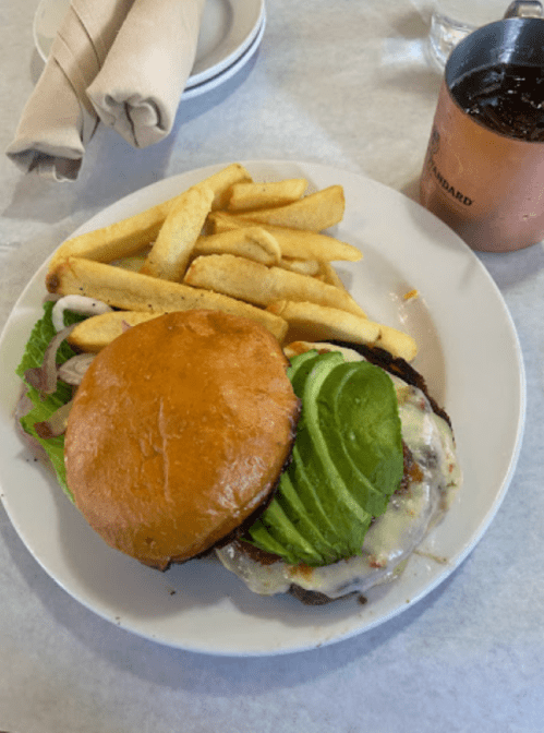 A plate with a burger topped with avocado, melted cheese, lettuce, and fries on the side.