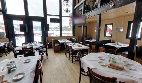 Interior of a cozy restaurant with tables set for dining, large windows, and a warm wooden decor.