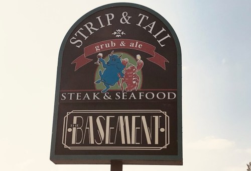 Sign for "Strip & Tail" featuring a blue crab and lobster, advertising steak and seafood, with "The Basement" below.