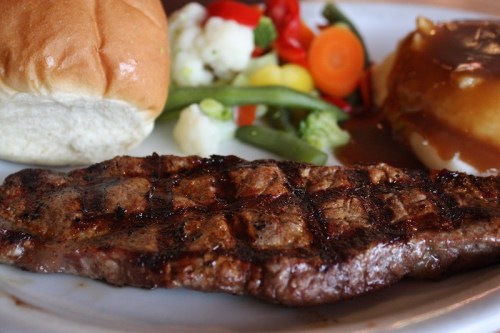 A grilled steak with a side of mixed vegetables, a dinner roll, and mashed potatoes with gravy on a plate.