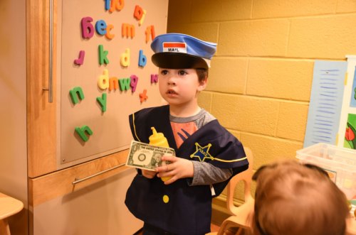 A young child in a mail carrier costume holds a dollar bill, standing in a colorful classroom with alphabet letters.