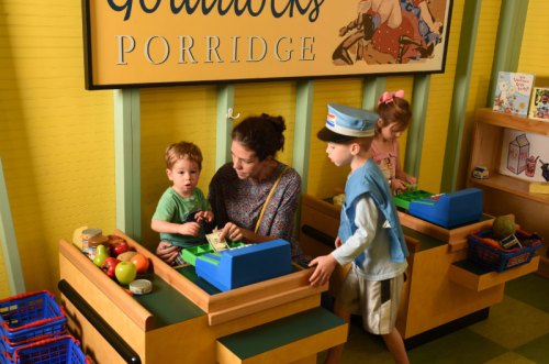 Children play at a pretend food market, interacting with a woman and using toy cash registers. Bright, colorful setting.