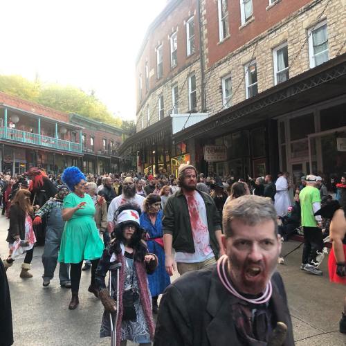 A crowd of people in costumes, including zombies and colorful outfits, walking down a historic street during a festival.