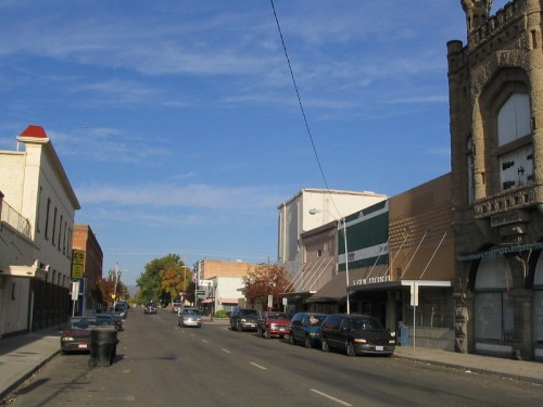 A quiet street scene featuring parked cars, storefronts, and a historic building under a clear blue sky.