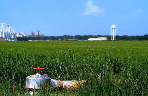 A gas cylinder lies in a green field, with a water tower and industrial buildings in the background under a clear blue sky.