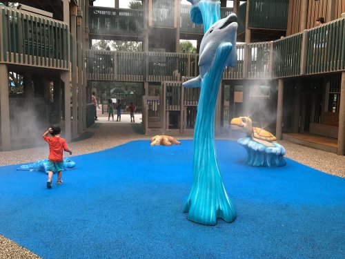 A child plays in a colorful splash pad with water features, including a dolphin and a turtle, surrounded by mist.