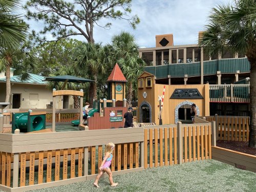 A colorful playground with various structures, surrounded by palm trees and a wooden fence, with children playing.