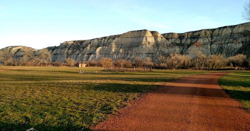 A scenic view of a grassy area with a dirt path leading to a large, rocky cliff in the background under a clear sky.