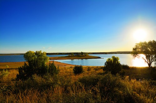 A serene landscape featuring a lake, grassy shore, and trees under a bright blue sky with a setting sun.