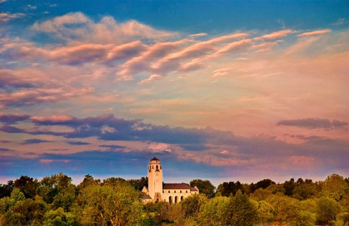 A scenic view of a clock tower surrounded by lush greenery under a colorful sky at sunset.
