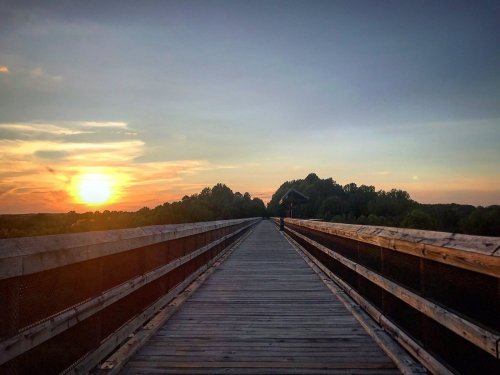 A wooden bridge stretches into the distance, framed by trees, with a vibrant sunset illuminating the sky.