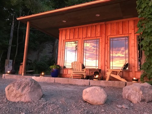 A cozy wooden cabin with large windows reflecting a colorful sunset, surrounded by rocks and greenery.