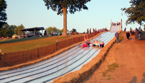 Children and families enjoy a large slide on a grassy hill, with spectators in the background during sunset.