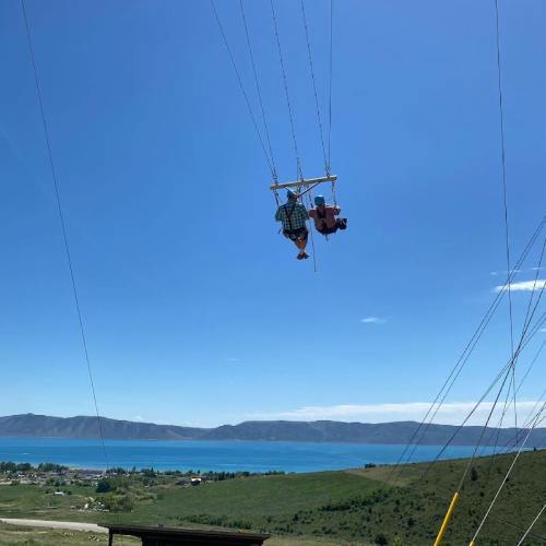 Two people are suspended in a harness, gliding over a scenic landscape with a lake and mountains in the background.