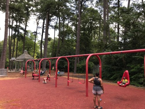 A playground with red swings and children playing, surrounded by tall trees and a gazebo in the background.