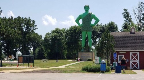 A large green statue of a man in a tunic stands in a park, surrounded by trees and a red barn in the background.