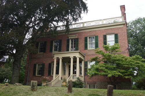 A historic brick house with green shutters, a porch supported by columns, and a rooftop terrace surrounded by trees.