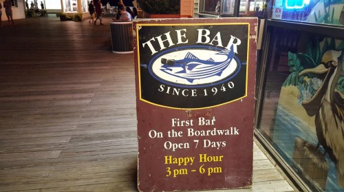 Sign for "The Bar" on the boardwalk, established in 1940, promoting happy hour from 3 PM to 6 PM. Open daily.