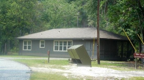 A gray building surrounded by trees, with a gravel road in front and a sign indicating the area.