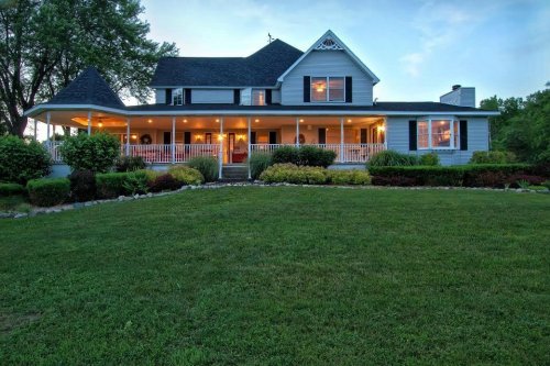 A charming two-story house with a wraparound porch, surrounded by lush greenery and illuminated at dusk.
