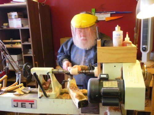 An elderly man with a beard works on a wood lathe, wearing a safety helmet and surrounded by woodworking tools.