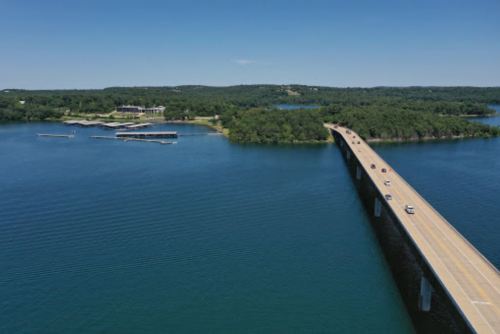 Aerial view of a bridge over a blue lake, surrounded by green trees and a small dock in the distance.