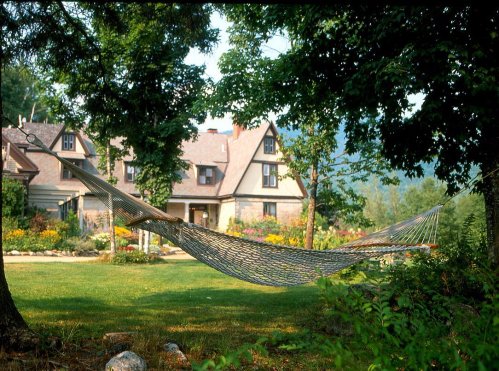 A hammock hangs between two trees in a lush garden, with a charming house and mountains in the background.