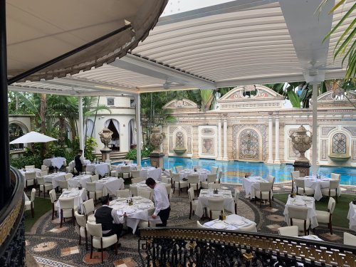 Elegant restaurant with outdoor seating, lush greenery, and a decorative pool in the background. Staff preparing tables.