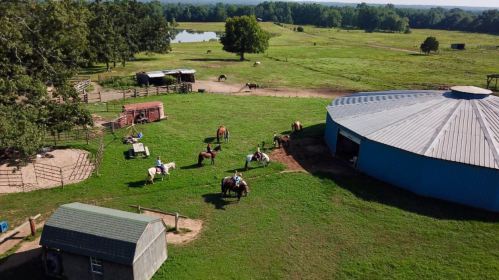 Aerial view of a farm with horses, a round barn, and a pond surrounded by green fields and trees.