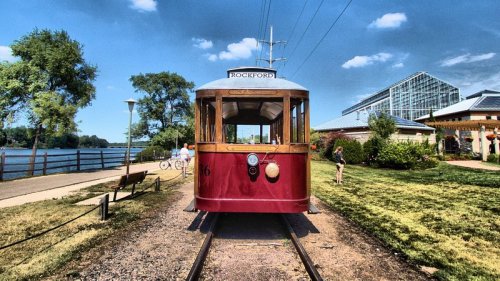 A red vintage tram on tracks by a river, with greenery and a glass building in the background under a blue sky.