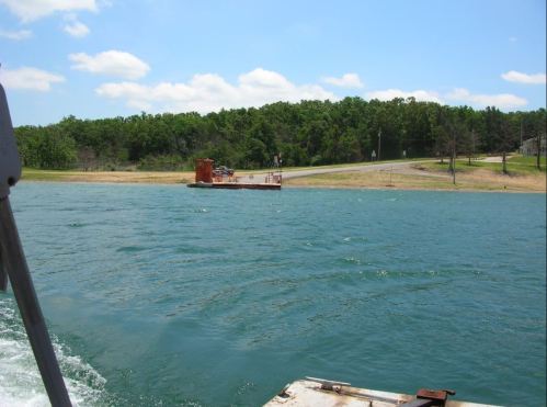A calm lake with a small ferry docked on the shore, surrounded by green trees and a clear blue sky.
