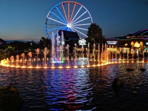 A colorful fountain lights up a pond at dusk, with a Ferris wheel in the background.