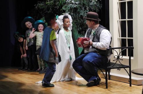 A group of children in costumes performs on stage, with one child in a white dress and another playing an accordion.