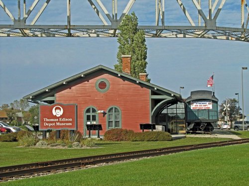 Red train depot museum with a sign, surrounded by green grass and a railway track, under a large bridge.