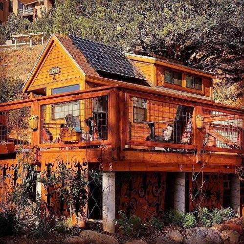 A cozy wooden cabin with a solar panel, surrounded by nature and featuring a deck with seating.
