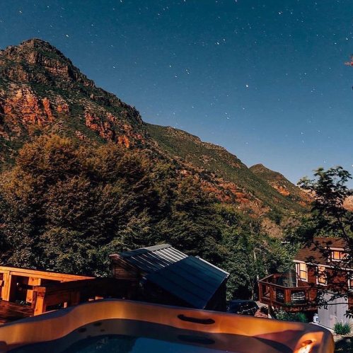 A serene night scene featuring mountains under a starry sky, with a hot tub in the foreground and trees nearby.