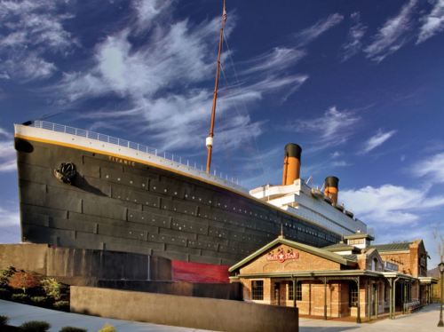 A large replica of the Titanic stands beside a historic building under a blue sky with clouds.