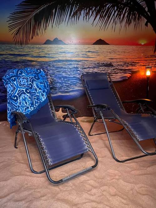 Two lounge chairs on sandy ground, with a blue patterned blanket draped over one, set against a beach sunset backdrop.