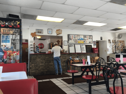 A casual diner interior with red and white seating, a counter, and a customer ordering at the register.