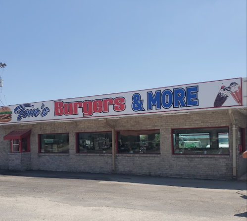 A restaurant building with a sign reading "Tom's Burgers & More," featuring large windows and a clear blue sky.