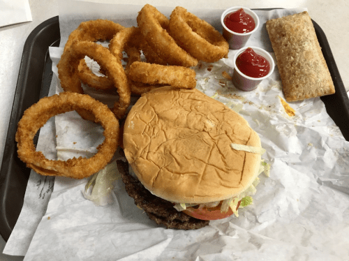 A tray with a burger, onion rings, two ketchup cups, and a pastry on a paper-lined surface.