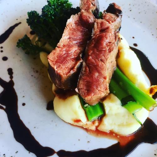 A plate of tender beef slices atop creamy mashed potatoes, garnished with green vegetables and a drizzle of sauce.